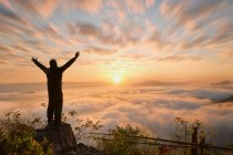 Silhouette d'un homme debout sur une montagne les bras tendus au lever du soleil, Thaïlande — Photo de stock