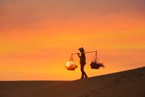 Mujer llevando cestas a través de una duna de arena al atardecer, Mui Ne, Vietnam - foto de stock