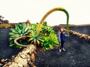 Niña de pie en un campo de lava junto a una planta gigante, Lanzarote, Islas Canarias, España - foto de stock