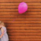 Chica de pie junto a una pared de madera sosteniendo un globo rosa - foto de stock