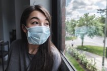 Woman wearing a face mask looking out of a window during lockdown — Stock Photo