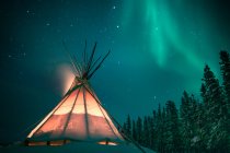 Prise de vue à longue exposition du tipi lumineux dans la forêt enneigée sous les aurores boréales, Yellowknife, Territoires du Nord-Ouest, Canada — Photo de stock
