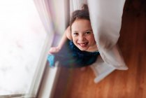 Portrait of a young girl standing by a window holding the curtain — Stock Photo