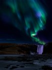 Uomo in piedi con le braccia tese sotto l'aurora boreale sopra Skogafoss, Islanda centro-meridionale, Islanda — Foto stock