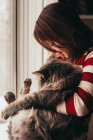 Улыбающаяся девушка, стоящая у окна и обнимающая кошку — стоковое фото
