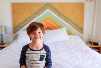 Rapaz sorridente sentado à beira de uma cama — Fotografia de Stock