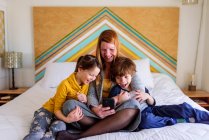 Mutter und Kinder lachen und schauen gemeinsam aufs Smartphone im Bett — Stockfoto
