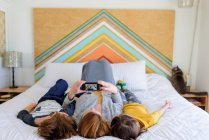Mutter und Kinder liegen zusammen im Bett und schauen aufs Smartphone — Stockfoto