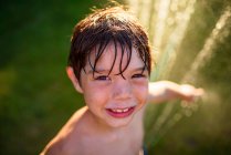 Porträt eines lächelnden Jungen, der im Garten an einem Wassersprenger steht, USA — Stockfoto