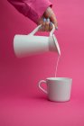 Arm outstretched holding ceramic jug and pouring milk at cup on pink background — Stock Photo