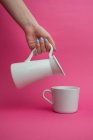Arm outstretched holding ceramic jug and pouring milk at cup on pink background — Stock Photo