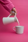 Arm ausgestreckt hält Keramikkrug und gießt Milch auf Tasse auf rosa Hintergrund — Stockfoto