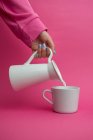 Braço esticado segurando jarro de cerâmica e derramando leite na xícara no fundo rosa — Fotografia de Stock