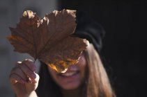 Adolescente sonriente sosteniendo una hoja de otoño frente a su cara, Argentina - foto de stock