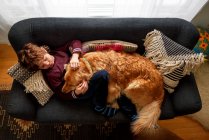 Boy lying on couch cuddling golden retriever dog — Stock Photo