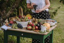 Mujer de pie junto a una mesa con frutas y verduras sosteniendo un ramo de uvas, Serbia - foto de stock