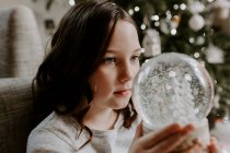 Menina sentada ao lado de uma árvore de Natal olhando para um globo de neve — Fotografia de Stock