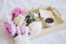 Bouquet de pivoines et une tasse de café avec une enveloppe sur un plateau sur un lit — Photo de stock
