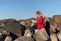 Жінка сидить на скелі біля моря і читає книжку 