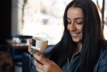 Жінка насолоджується чашкою чаю, використовуючи свій мобільний телефон — стокове фото