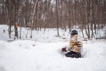 Мальчик в зимней одежде, покрытый снегом, играет со снегом в парке — стоковое фото