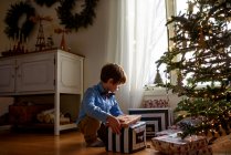 Niño arrodillado frente a un árbol de Navidad mirando regalos - foto de stock