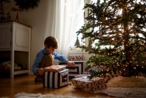Menino ajoelhado na frente de uma árvore de Natal olhando para presentes — Fotografia de Stock