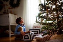 Niño arrodillado frente a un árbol de Navidad con regalos mirando hacia arriba - foto de stock
