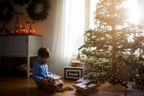 Menino sentado na frente de uma árvore de Natal segurando presentes — Fotografia de Stock