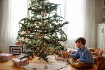 Junge sitzt am Weihnachtsbaum und betrachtet Dekorationen — Stockfoto