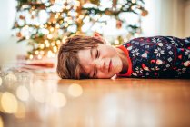 Niño tirado en el suelo delante de un árbol de Navidad tirando caras raras - foto de stock