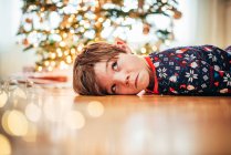 Ragazzo sdraiato sul pavimento davanti a un albero di Natale che tira facce strane — Foto stock