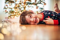 Menino deitado no chão em frente a uma árvore de Natal rindo — Fotografia de Stock