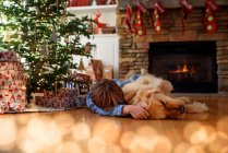 Menino deitado no chão em frente a uma árvore de Natal abraçando seu cão — Fotografia de Stock