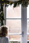 Ragazza che guarda la neve da una finestra decorata con abeti e luci fatate a Natale — Foto stock