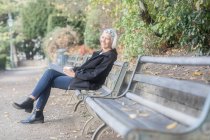 Mulher sorridente sênior sentado no banco do parque com xícara de café — Fotografia de Stock