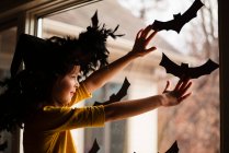 Chica sonriente con un sombrero de brujas pegando decoraciones de murciélagos en una ventana, Estados Unidos - foto de stock