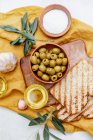 Green olives with olive oil with fresh baked ciabatta bread,  salt and young olive branches over white background. Flat lay, space — Stock Photo