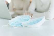 El primer plano de dos máscaras faciales en una mesa y dos mujeres trabajando en un portátil en el fondo - foto de stock