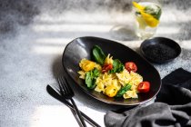 Салат с фарфоровой пастой и овощами подается в черной керамической миске со стаканом лимона и базиликовой водой — стоковое фото
