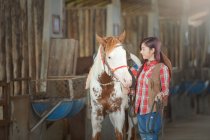 Woman standing in a stable with her horse ready to go riding — Stock Photo