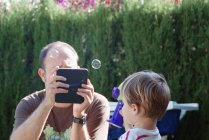 Pai tirando uma foto de seu filho segurando uma máquina de varinha de bolha com um tablet digital — Fotografia de Stock