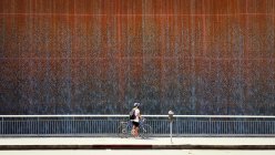 Teenage boy standing with his bicycle on pavement by a waterfall on a wall, USA — Stock Photo