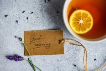 Taza de té de limón, flores de lavanda y etiqueta de buenos días en el fondo de hormigón - foto de stock