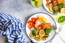 Two servings of grilled salmon steak with roasted courgettes, tomatoes, lemon and a glass of lemon water — Stock Photo