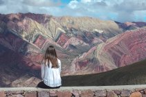 Rückansicht einer Frau, die an einer Wand sitzt und auf die Berglandschaft blickt, El Hornacal, Jujuy, Argentinien — Stockfoto