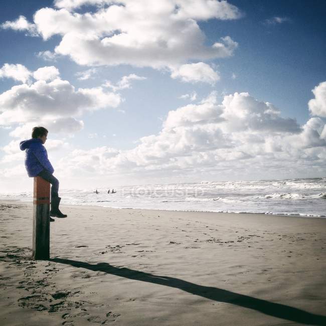 Menino sentado em poste de madeira na praia — Fotografia de Stock