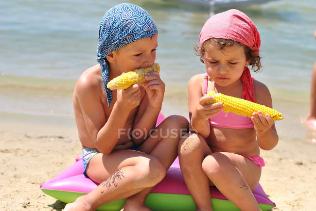 Girl and boy eating sweetcorn on beach — Stock Photo
