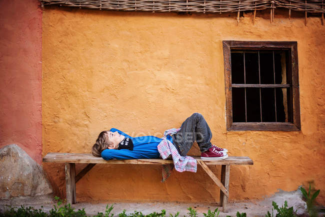 Menino deitado no banco perto da casa — Fotografia de Stock