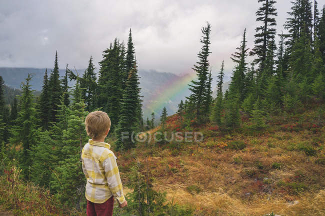 Menino de pé na montanha olhando para o arco-íris — Fotografia de Stock